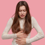 Image of unhappy woman suffers from stomachache after eating spoiled food, feels discomfort in belly, has disorder, wears casual clothes, models against pink background. Painful disease conccept