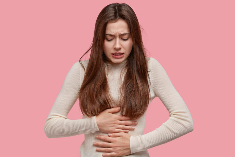 Image of unhappy woman suffers from stomachache after eating spoiled food, feels discomfort in belly, has disorder, wears casual clothes, models against pink background. Painful disease conccept